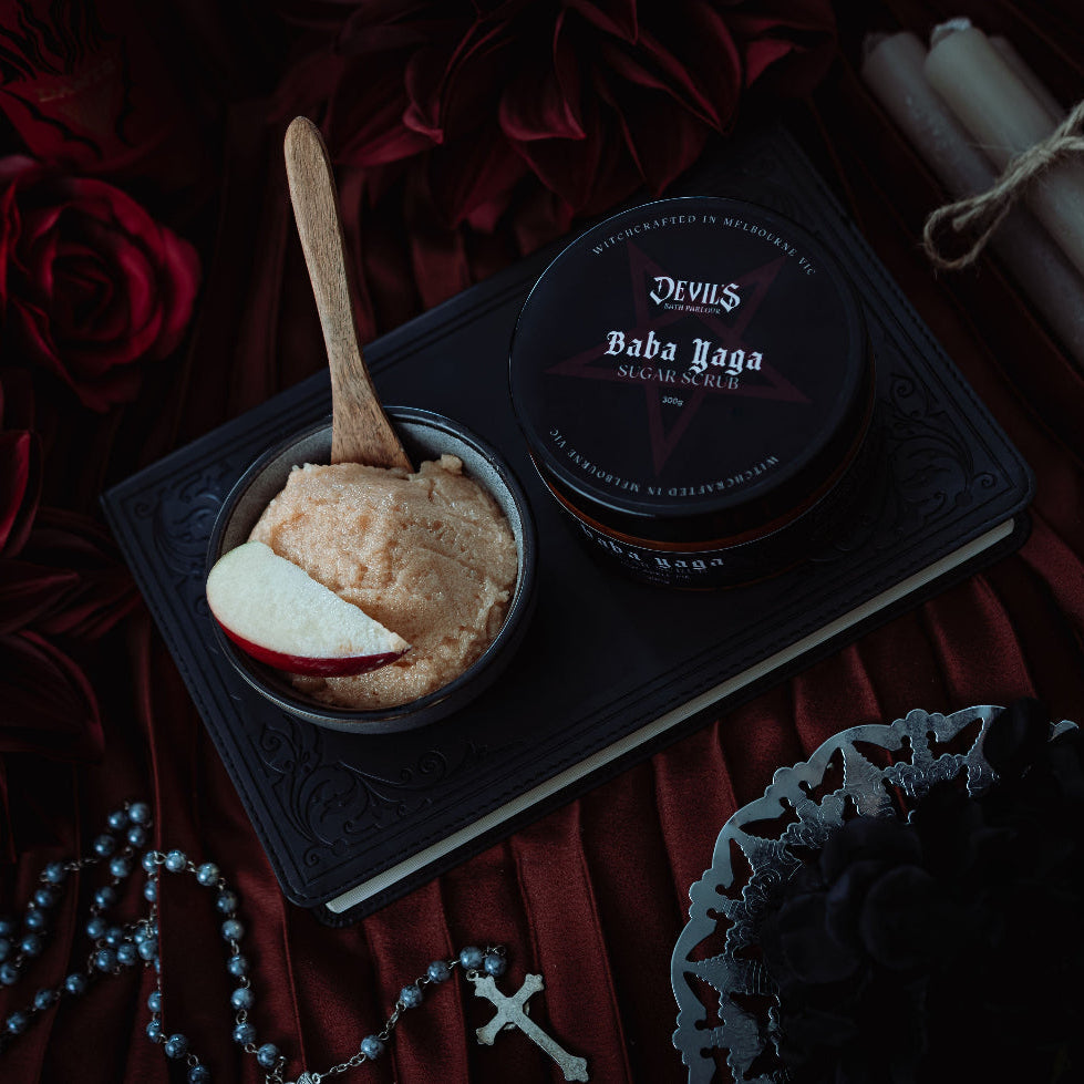 A styled flat-lay featuring a jar of Devil's Baba Yaga ritual salt on a black book, next to a bowl of salt with a wooden spoon and apple slice, surrounded by dark red roses, candles, and a rosary.